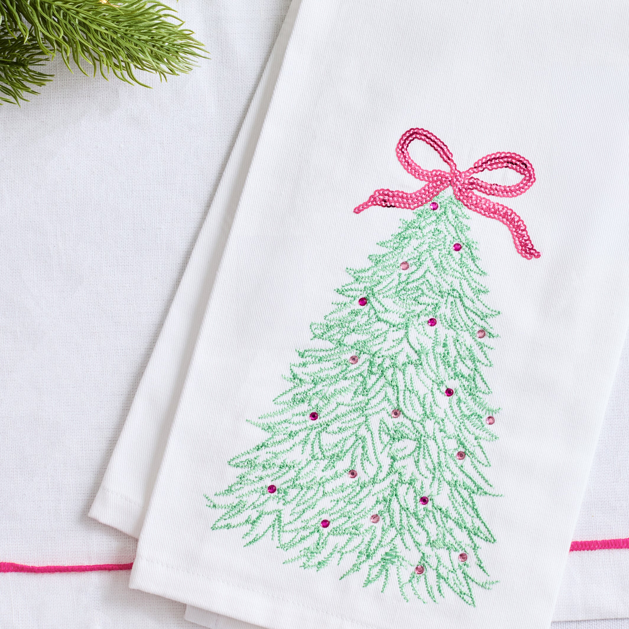 A folded white fabric napkin or tea towel featuring an embroidered green Christmas tree with a pink bow and small pink ornaments, styled on a white surface with a sprig of green pine in the corner.