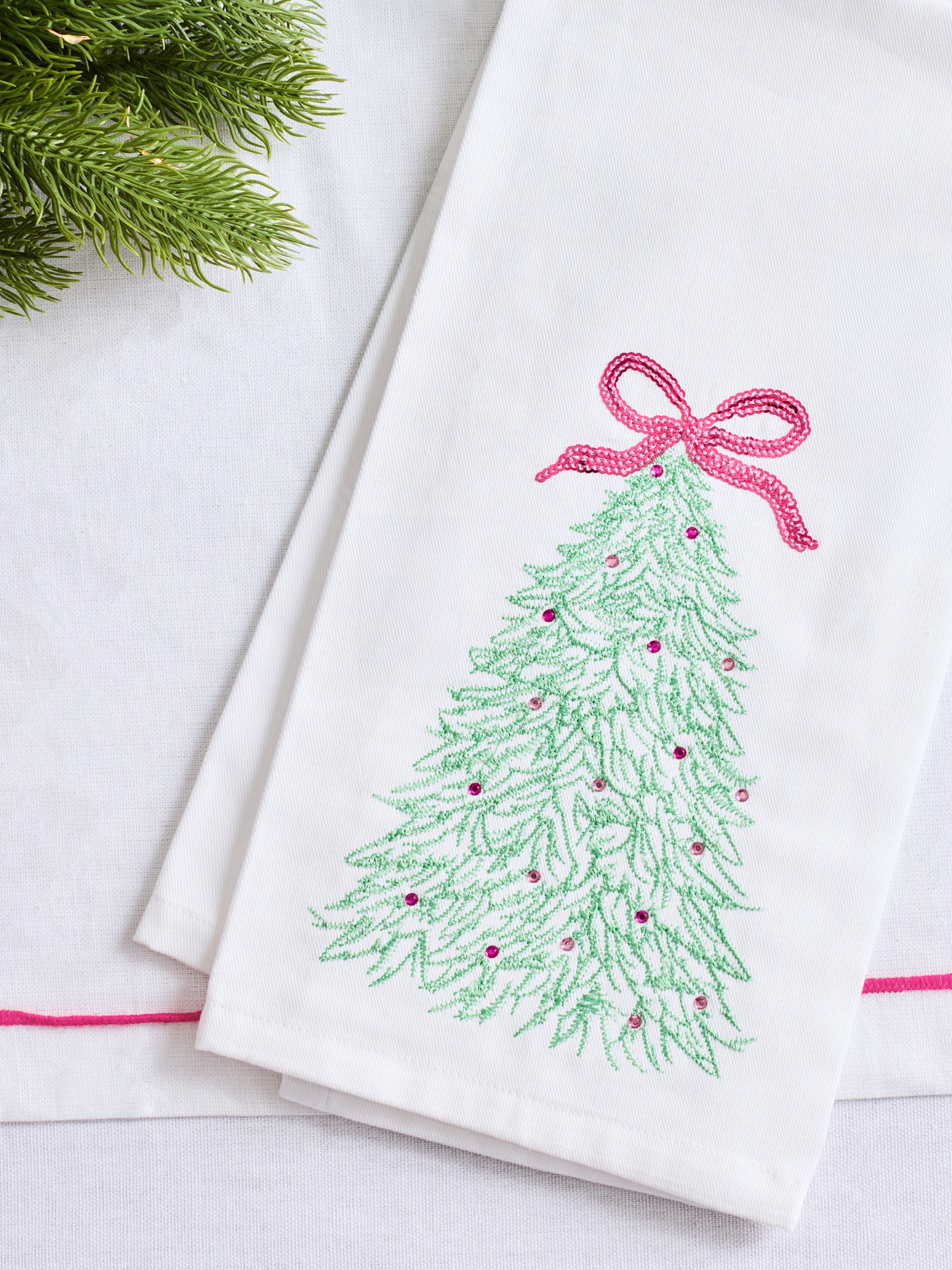 A folded white fabric napkin or tea towel featuring an embroidered green Christmas tree with a pink bow and small pink ornaments, styled on a white surface with a sprig of green pine in the corner.