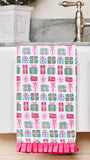 A white kitchen towel with a pink, green, and blue gift box pattern, draped over the edge of a white sink with a ruffled pink bottom hem.