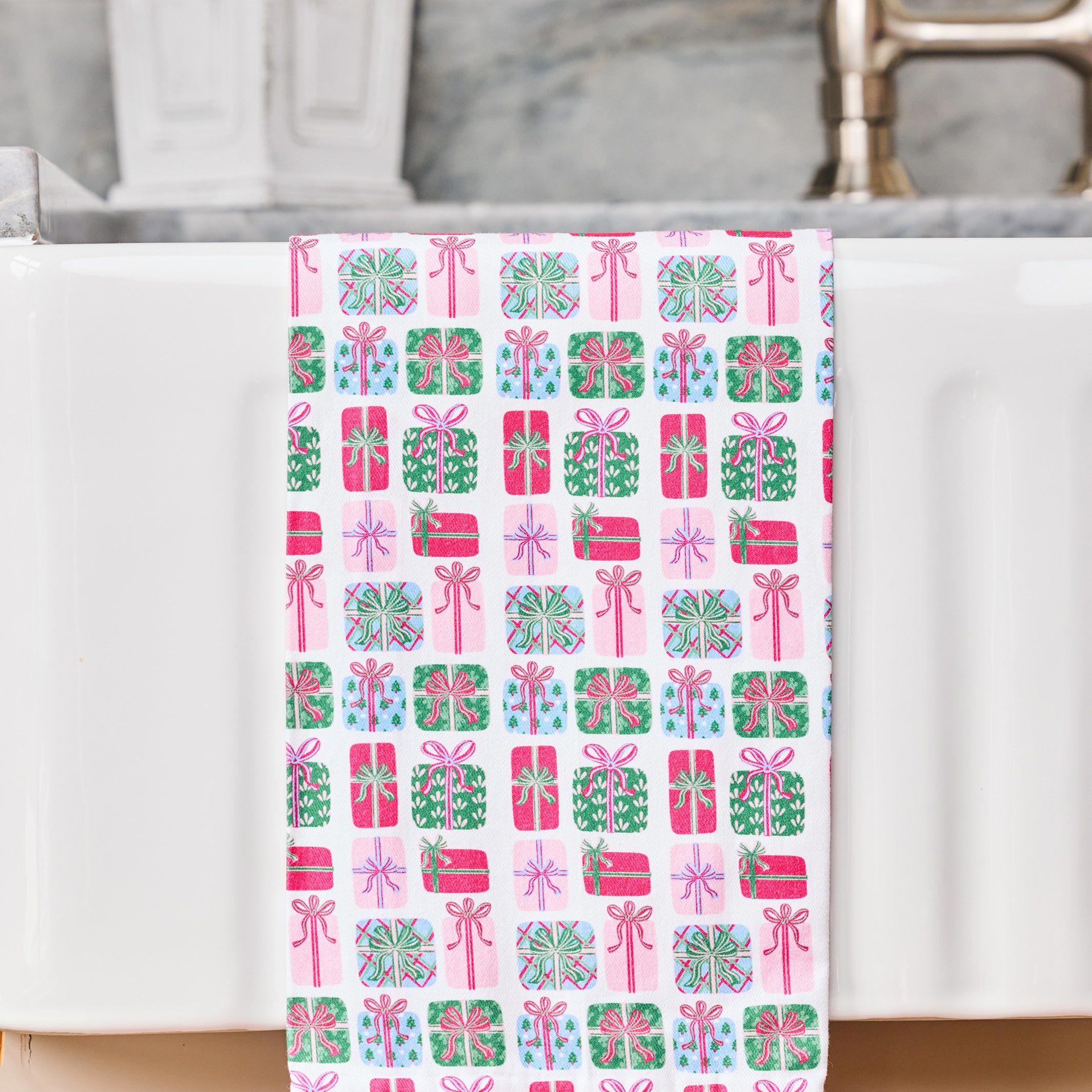 A white kitchen towel with a pink, green, and blue gift box pattern, draped over the edge of a white sink with a ruffled pink bottom hem.