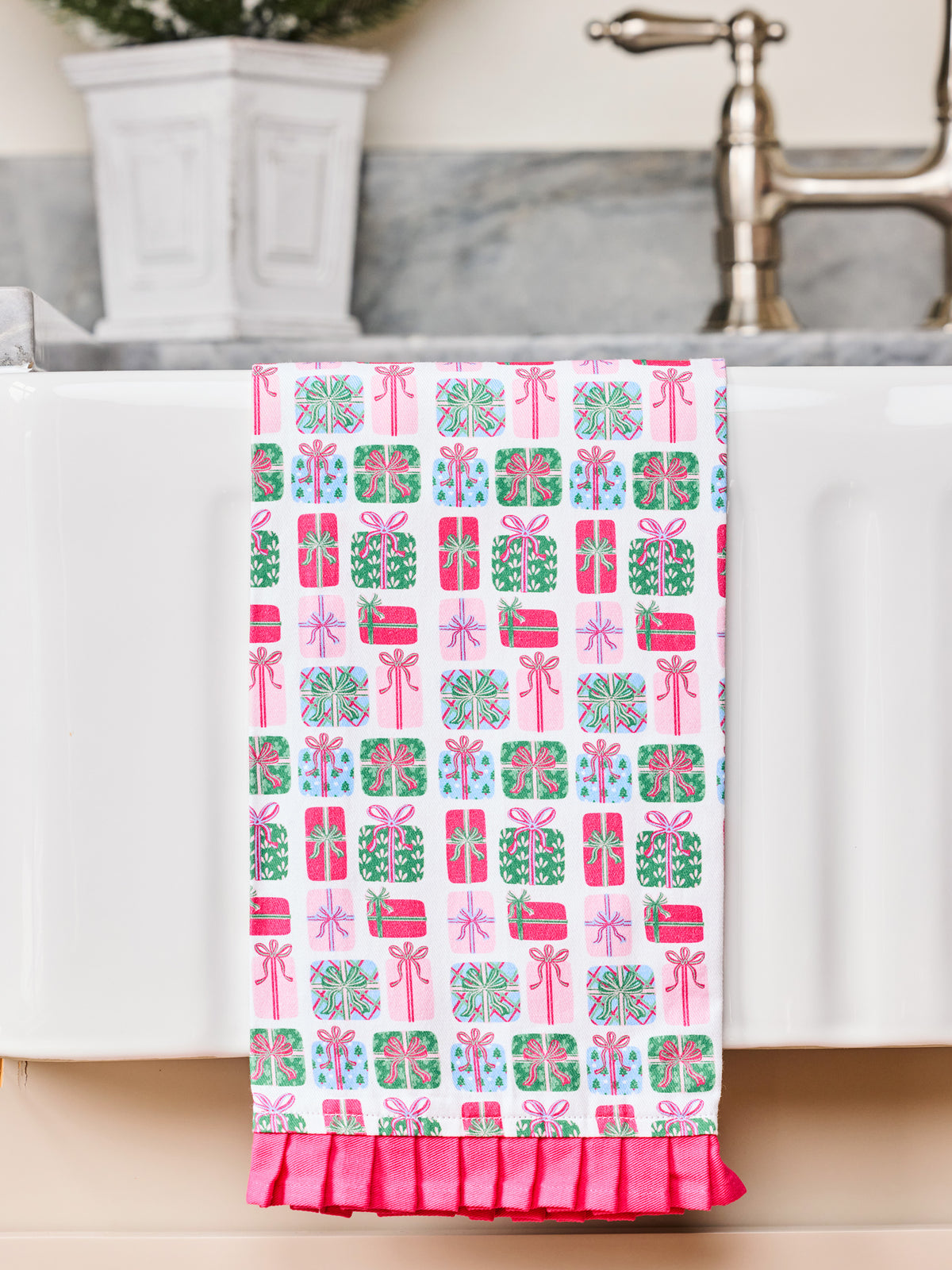 A white kitchen towel with a pink, green, and blue gift box pattern, draped over the edge of a white sink with a ruffled pink bottom hem.