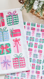 A white kitchen towel with a colorful Christmas gift pattern, displayed next to a matching gift-patterned sheet on a light beige surface with a wicker basket and evergreen branches in the background.