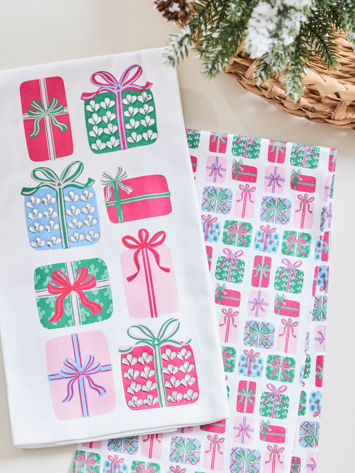A white kitchen towel with a colorful Christmas gift pattern, displayed next to a matching gift-patterned sheet on a light beige surface with a wicker basket and evergreen branches in the background.