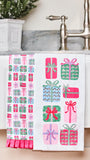 Two white kitchen towels with holiday gift patterns hanging over a white sink in a bathroom. One towel features a vertical column of pink, green, and blue wrapped presents, and the other features a grid of various wrapped presents in pink, green, and blue.