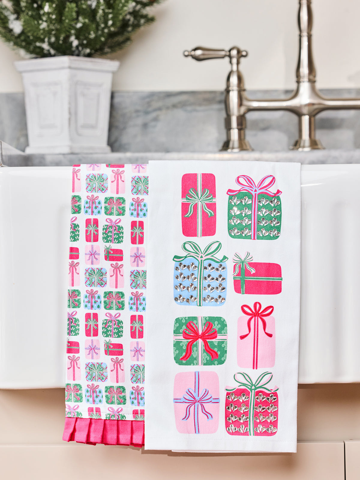 Two white kitchen towels with holiday gift patterns hanging over a white sink in a bathroom. One towel features a vertical column of pink, green, and blue wrapped presents, and the other features a grid of various wrapped presents in pink, green, and blue.