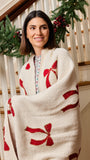 A woman wrapped in a cream-colored knit blanket with red bow patterns, standing on a decorated indoor staircase.