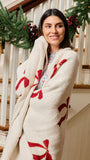 A woman wrapped in a cream-colored, plush knit blanket with red holiday motifs, standing in front of a decorated staircase with green garlands and red berries.