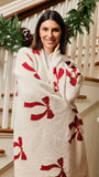 A woman wrapped in a cozy cream-colored blanket with red bow patterns, standing on a decorated indoor staircase.