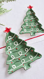 Two green Christmas tree-shaped ceramic dishes with white icing-like swirls and red star accents, displayed on a white surface with holiday greenery and a gold bell as props.