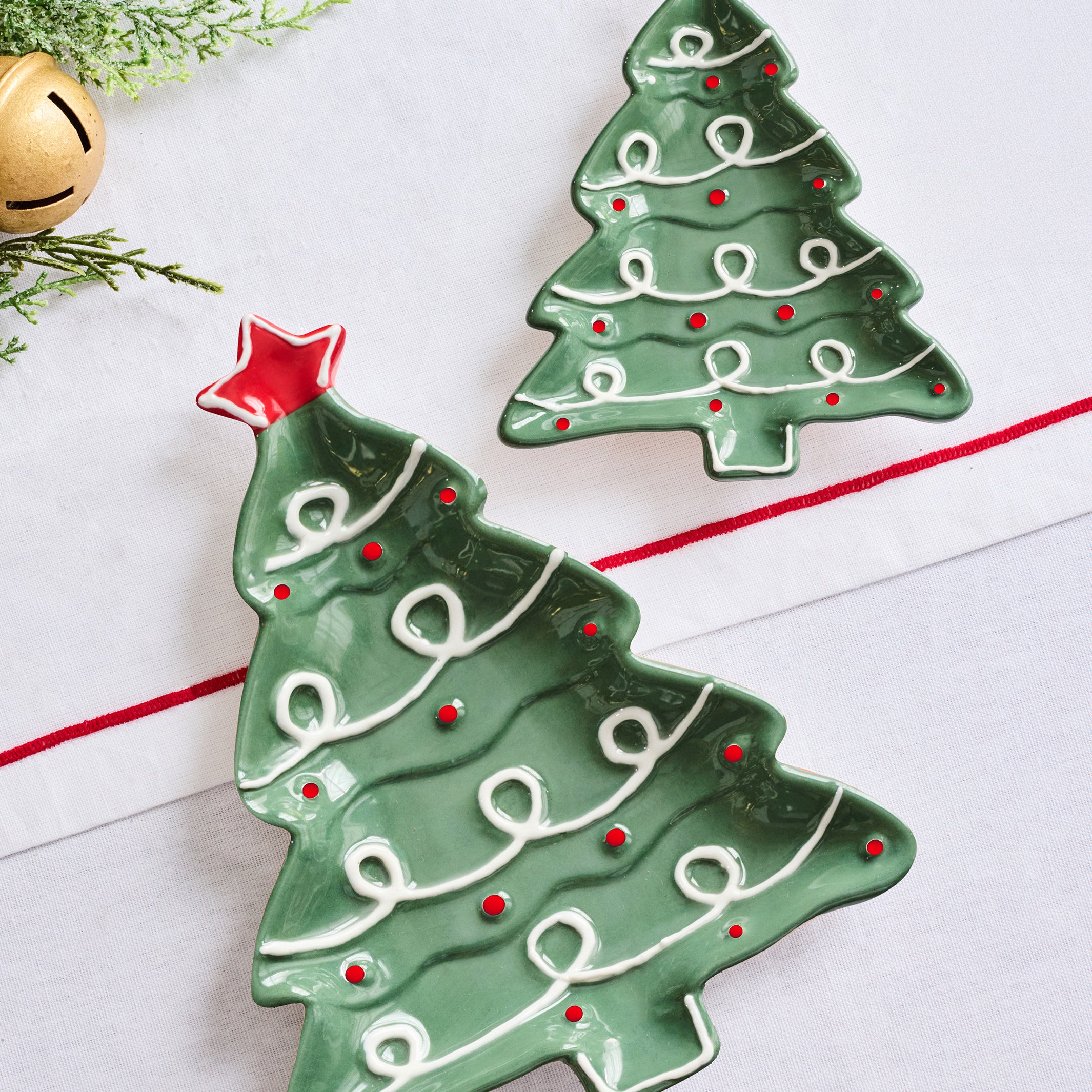 Two green Christmas tree-shaped ceramic dishes with white icing-like swirls and red star accents, displayed on a white surface with holiday greenery and a gold bell as props.