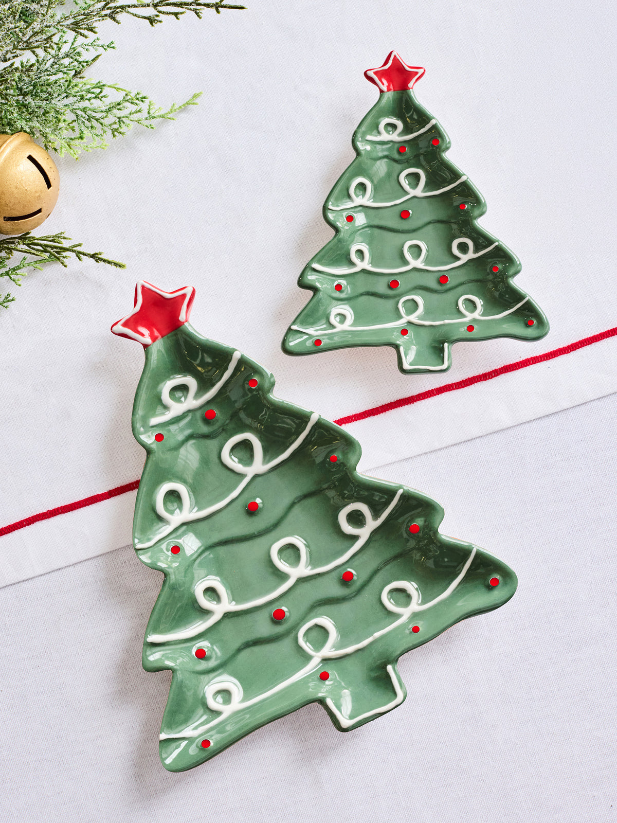 Two green Christmas tree-shaped ceramic dishes with white icing-like swirls and red star accents, displayed on a white surface with holiday greenery and a gold bell as props.