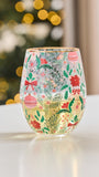 A clear stemless wine glass decorated with a festive Christmas pattern of red ornaments, green pine branches, red bows, and holly leaves, sitting on a white surface with a blurred holiday tree and warm lights in the background.