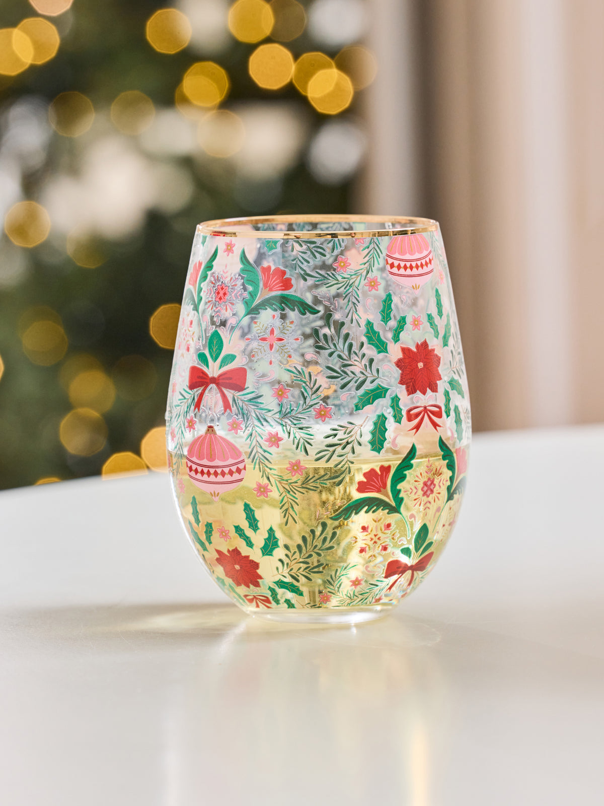 A clear stemless wine glass decorated with a festive Christmas pattern of red ornaments, green pine branches, red bows, and holly leaves, sitting on a white surface with a blurred holiday tree and warm lights in the background.