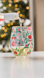 A festive stemless wine glass with a Christmas pattern, filled with white wine as a bottle pours liquid into it, set against a blurred holiday background.