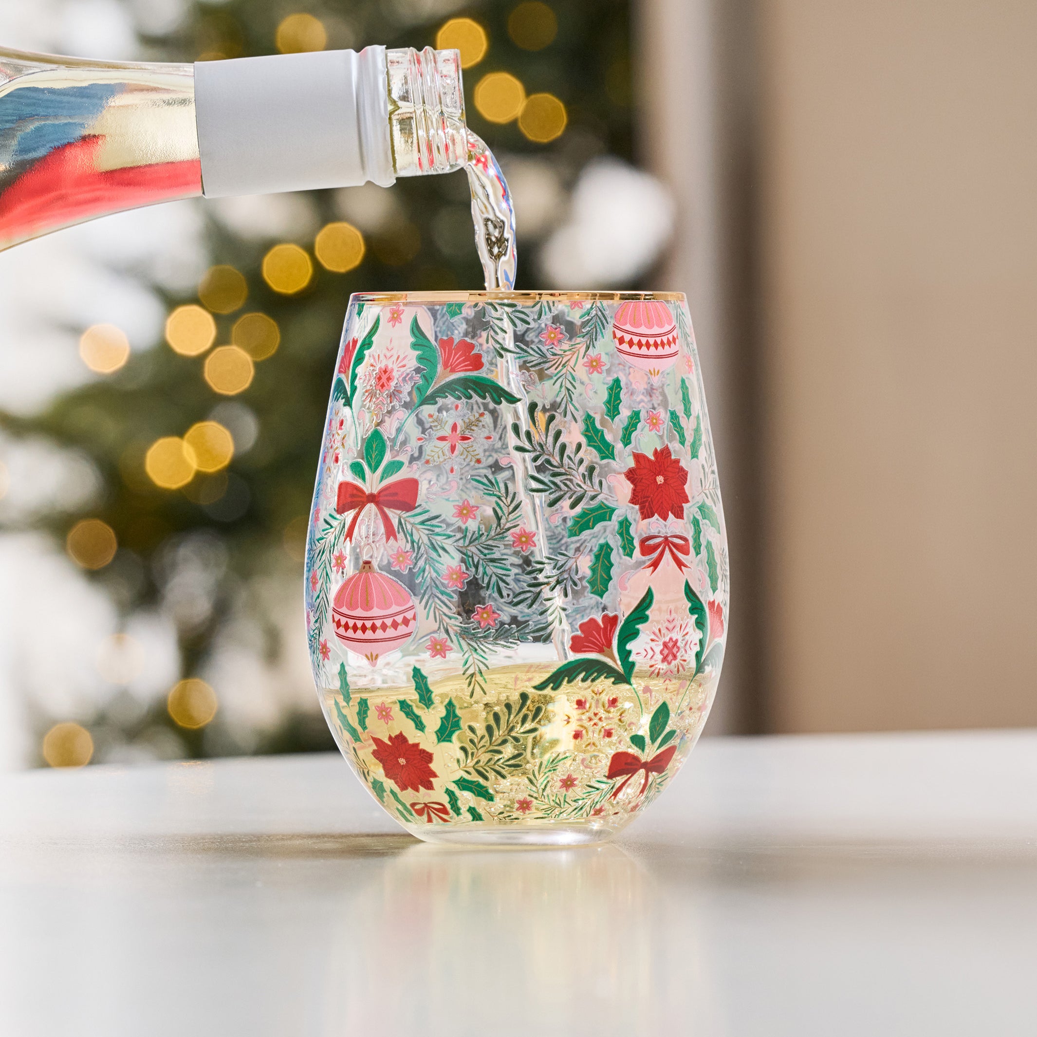 A festive stemless wine glass with a Christmas pattern, filled with white wine as a bottle pours liquid into it, set against a blurred holiday background.