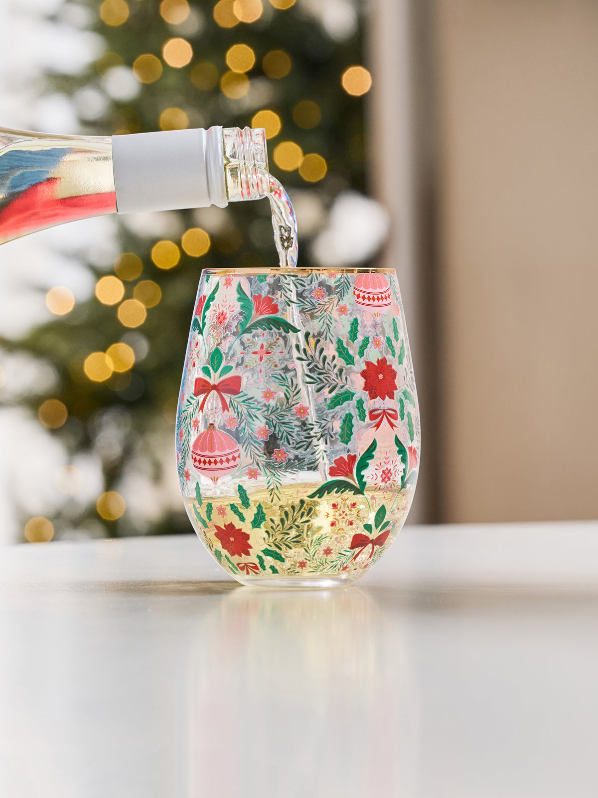 A festive stemless wine glass with a Christmas pattern, filled with white wine as a bottle pours liquid into it, set against a blurred holiday background.