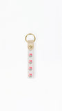 A white fabric keychain strap featuring a vertical pattern of pink roses with green leaves, finished with a beige leather tab and a gold-tone metal key ring at the top.