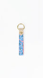 A vertical floral keychain strap with a blue, pink, and green pattern, finished with a beige leather tab and a gold-tone metal key ring.