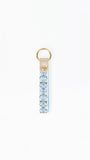 A vertical leather keychain strap featuring a light blue and white floral pattern, finished with a gold-tone metal ring and a small gold logo medallion on the leather tab.