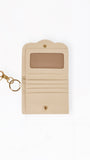 A beige leather card holder wallet with a gold-tone key ring and clasp, featuring a clear ID window on the front, shown on a plain white background.