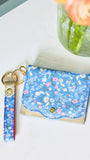 A blue floral card holder wallet with a matching fabric wristlet strap keychain, displayed on a white marble surface next to a glass vase with flowers.