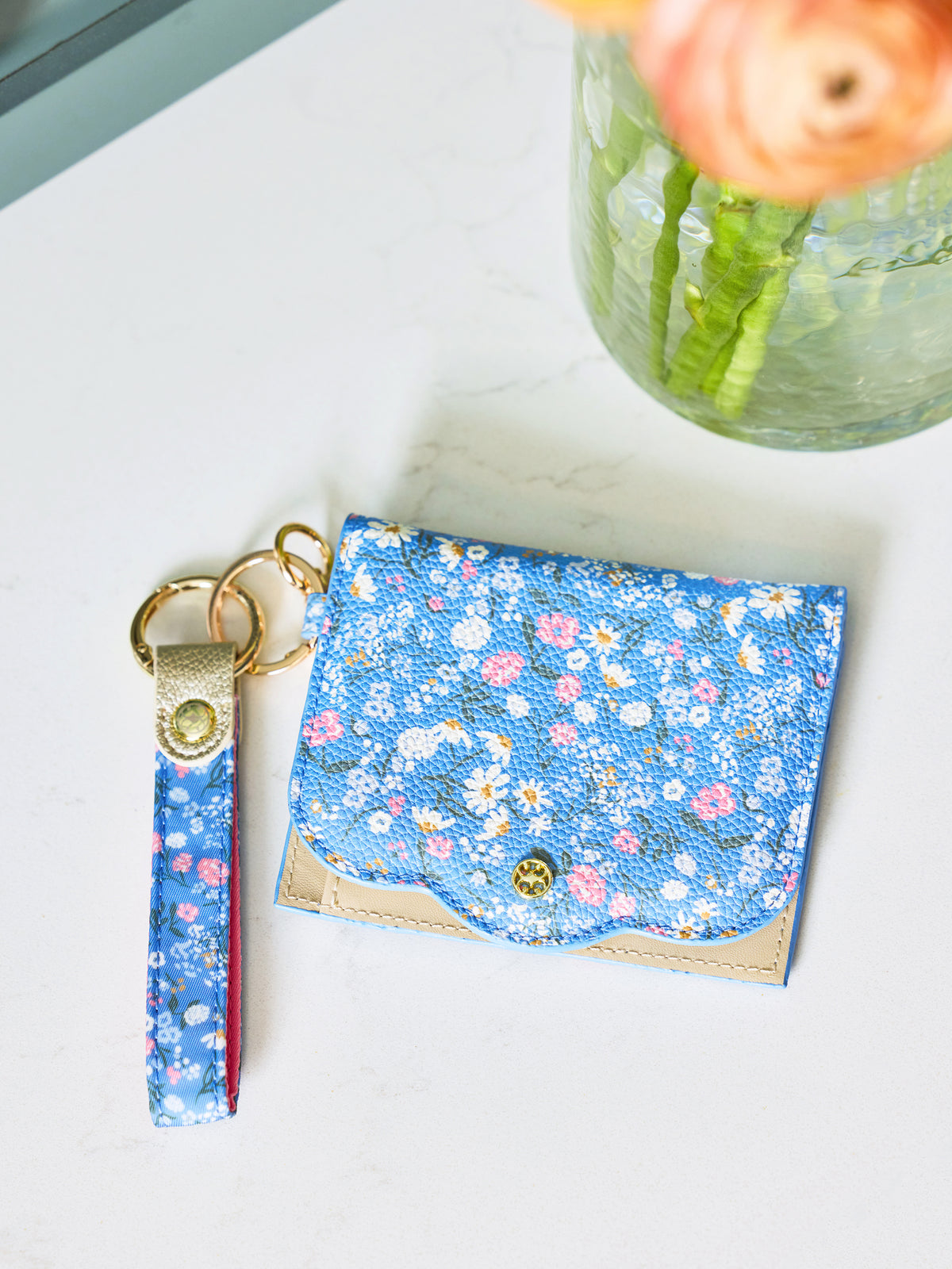 A blue floral card holder wallet with a matching fabric wristlet strap keychain, displayed on a white marble surface next to a glass vase with flowers.