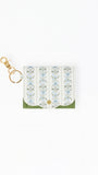 A small white leather card holder or wallet with a delicate blue and green floral pattern, a green bottom border, and a gold-tone key ring clasp on a plain white background.