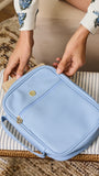 A light blue leather bible cover with gold-tone hardware, held by a person wearing a beige dress with blue floral embroidery, sitting on a striped cushion.