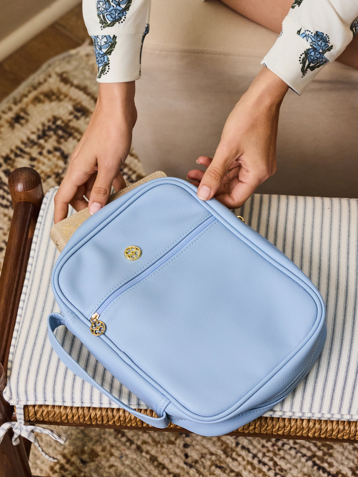 A light blue leather bible cover with gold-tone hardware, held by a person wearing a beige dress with blue floral embroidery, sitting on a striped cushion.