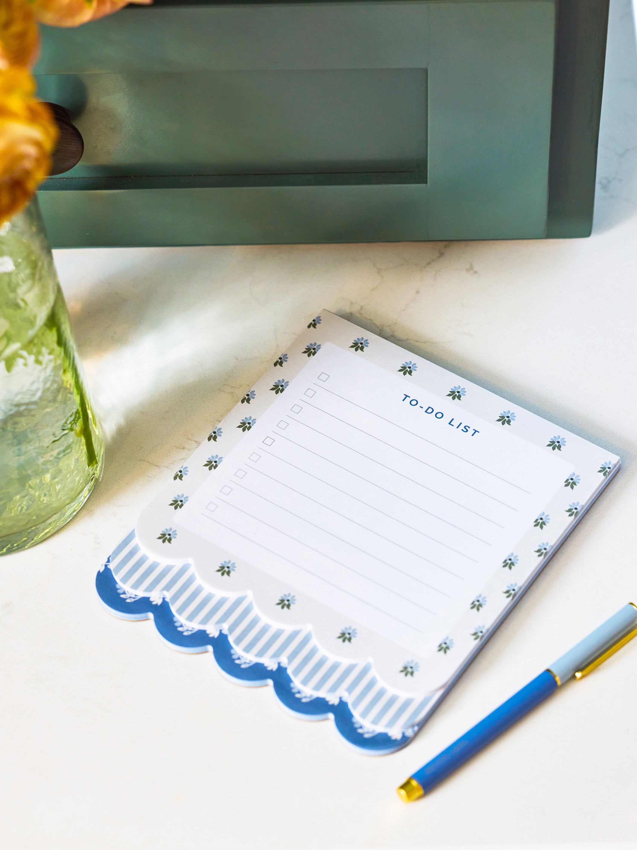 A white to-do list notepad with a blue scalloped border and small blue floral pattern, styled on a light marble surface next to a blue pen and a green glass vase.