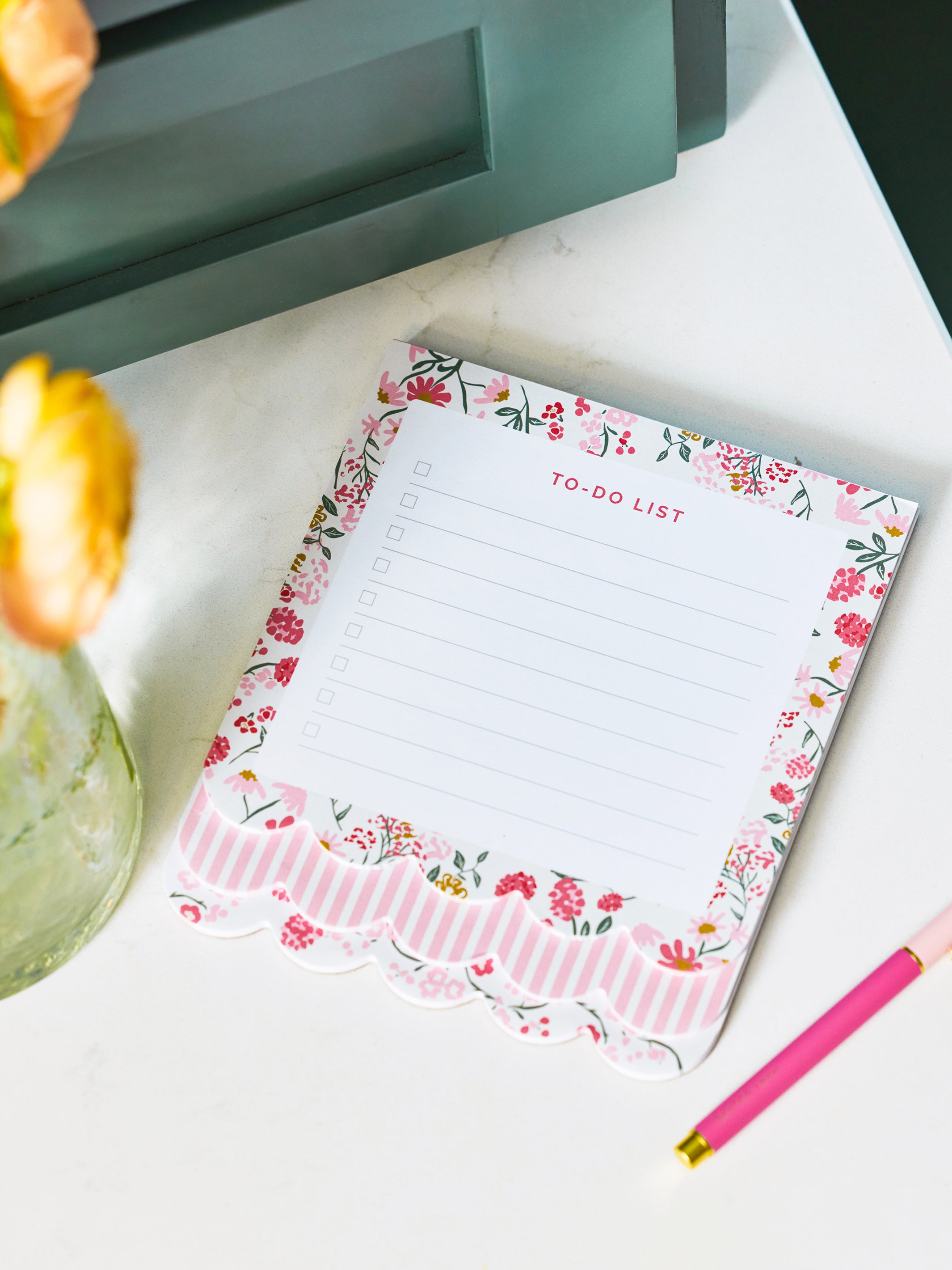 A floral and striped notepad with a scalloped edge, labeled 'TO-DO LIST', placed on a white surface with a pink pen nearby and blurred flowers in the background.