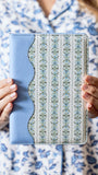 A person in a blue and white floral pajama shirt holds a tablet case with a light blue leather-like spine and a cream floral patterned back.