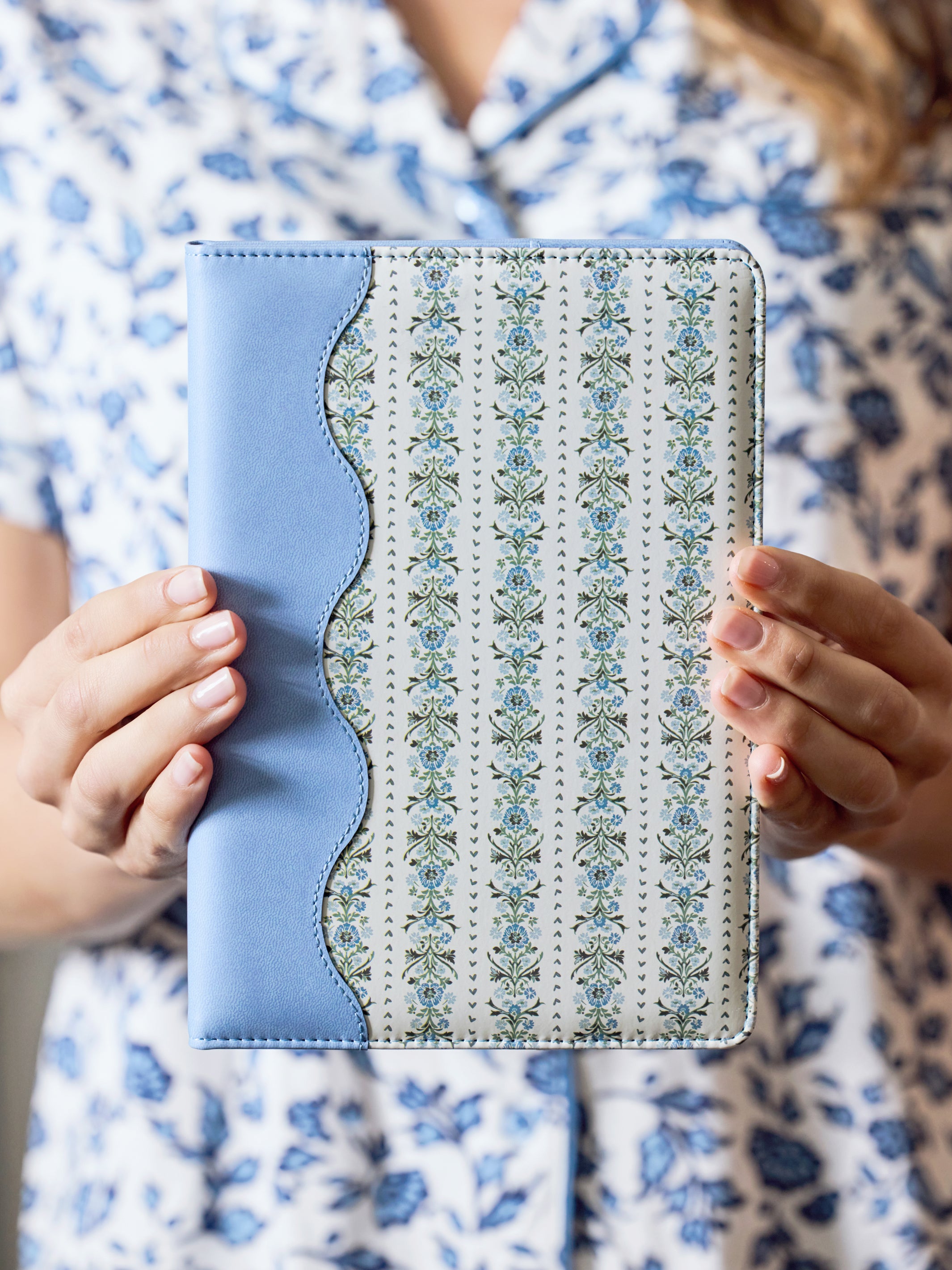 A person in a blue and white floral pajama shirt holds a tablet case with a light blue leather-like spine and a cream floral patterned back.