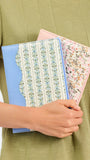 A person in an olive green dress holds two slim folio-style cases against their torso. The front case is light blue with a cream vertical stripe panel featuring blue floral motifs, and the back case is pink with a delicate multicolor floral print.