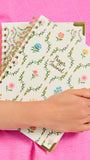 A hand holds a stack of spiral-bound prayer journals with floral illustrations and gold corner accents against a bright pink fabric background.