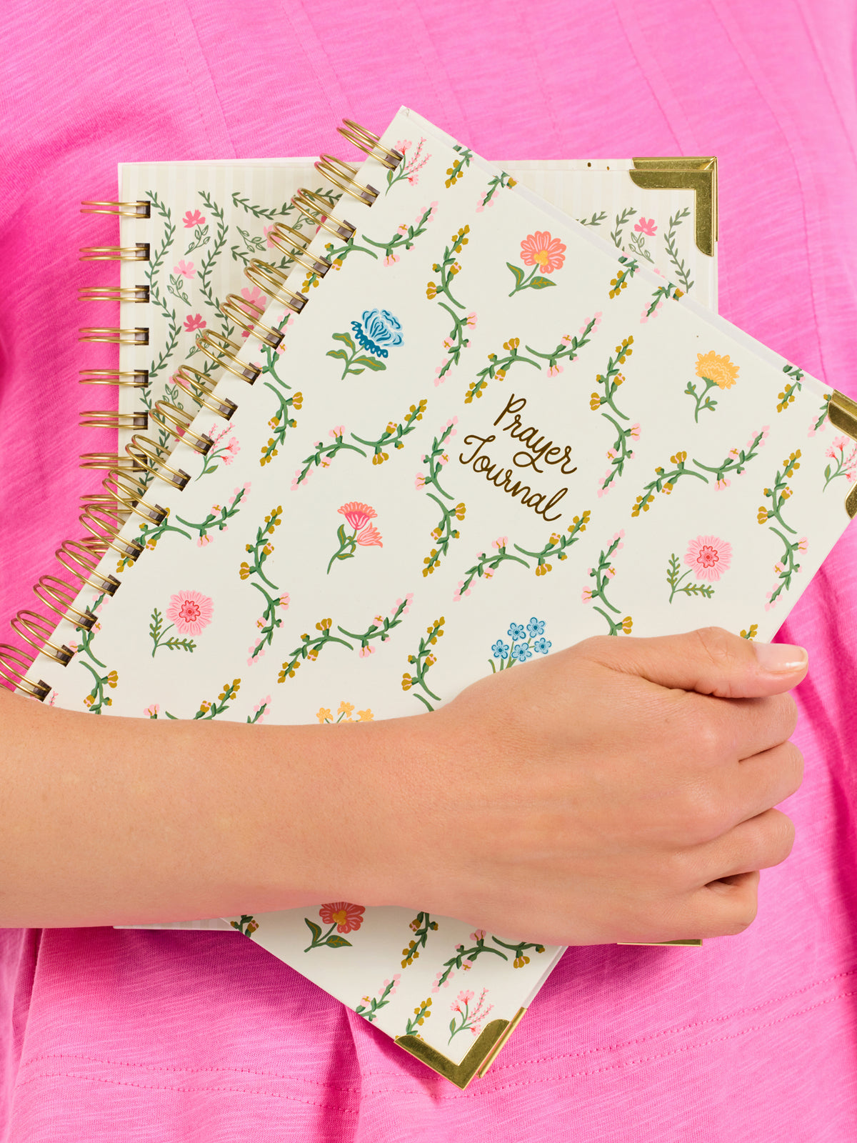 A hand holds a stack of spiral-bound prayer journals with floral illustrations and gold corner accents against a bright pink fabric background.