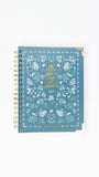 A spiral-bound notebook with a dusty blue cover decorated with white floral patterns and gold foil text reading 'with courage before me and hope behind me'.
