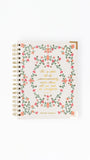 A spiral-bound prayer journal with a cream-colored cover decorated with a pink and green floral wreath and gold foil lettering, shown on a plain white background.