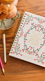 A spiral-bound journal with a white cover decorated with a pink and green floral border and gold script text, placed on a wooden table next to two pens and a vase of peach roses.