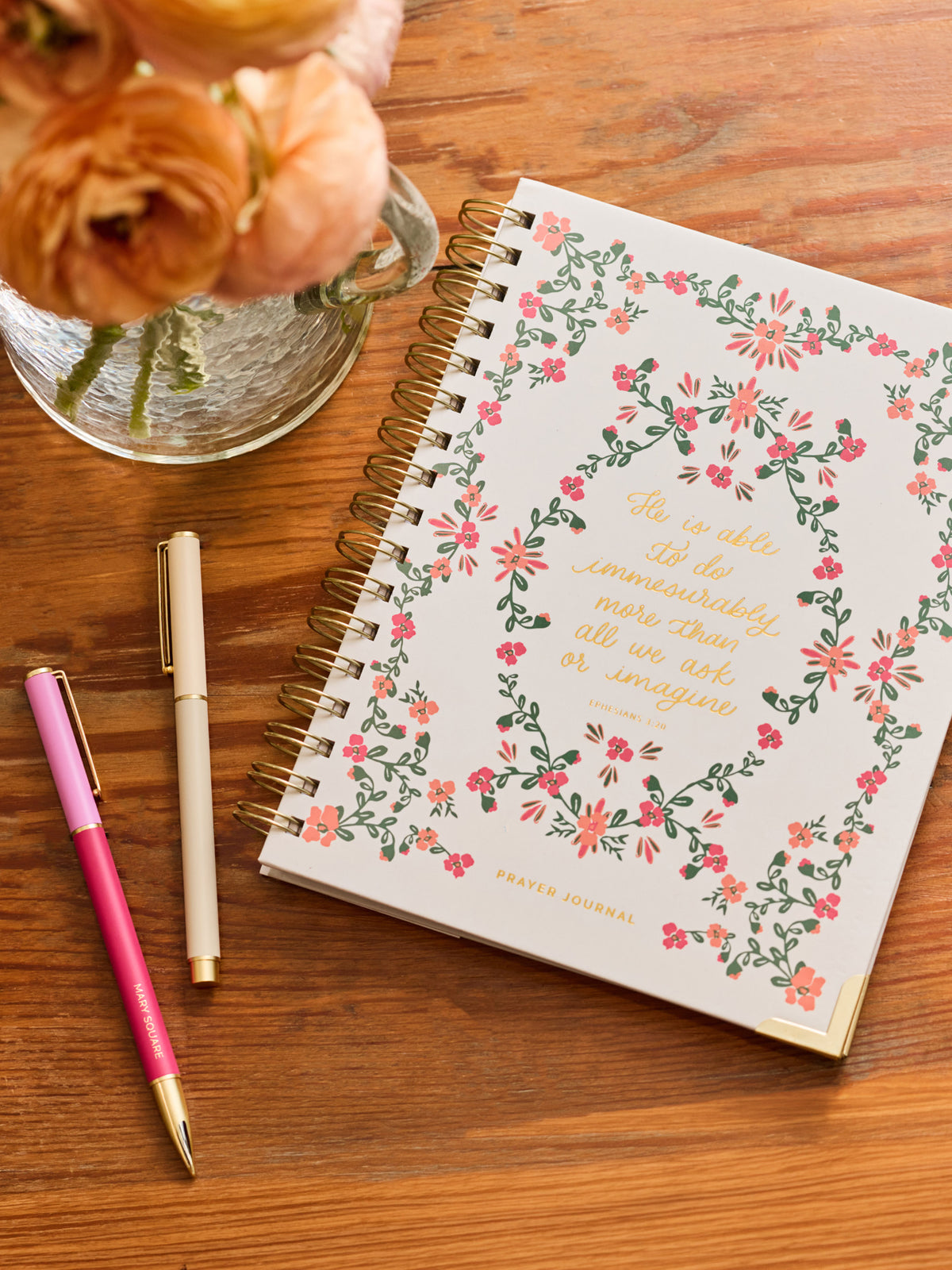 A spiral-bound journal with a white cover decorated with a pink and green floral border and gold script text, placed on a wooden table next to two pens and a vase of peach roses.