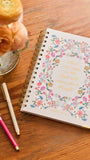 A spiral-bound journal with a floral border and gold lettering reading 'those who hope in the Lord will know their strength' lies on a wooden table beside two pens and a vase of peach flowers.