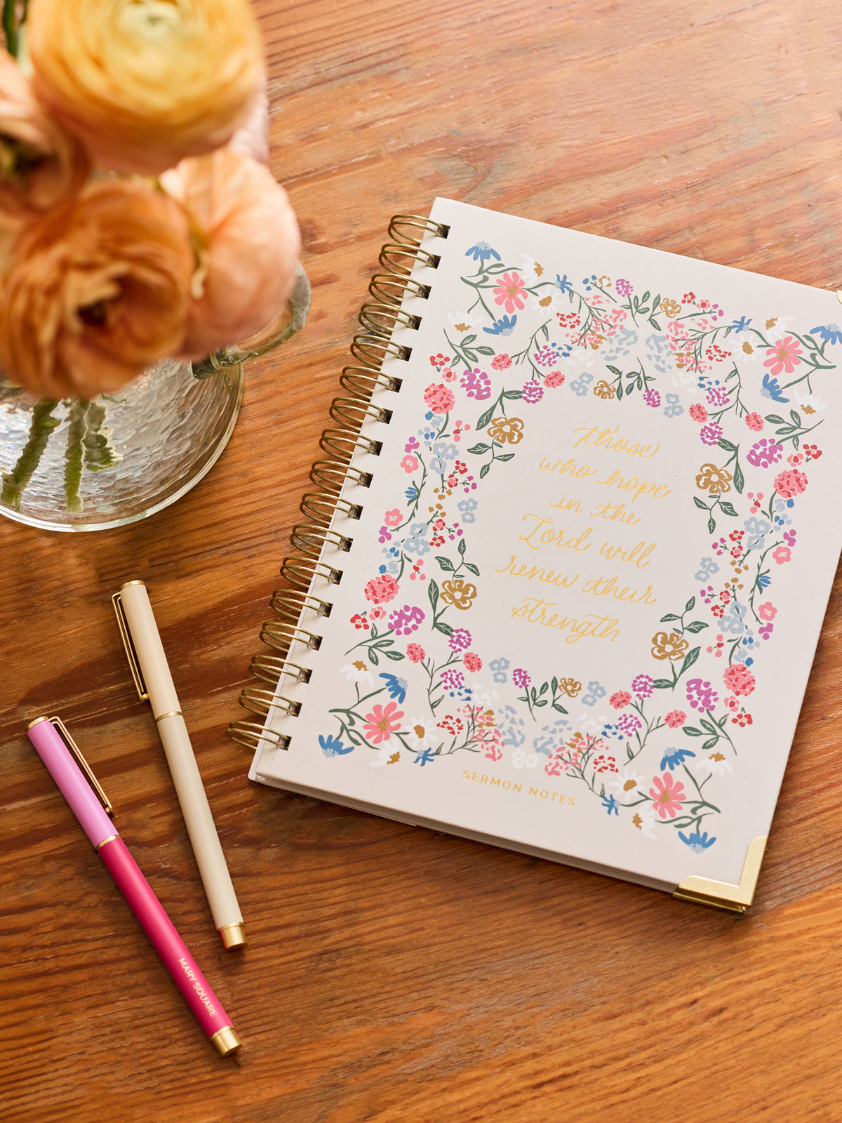 A spiral-bound journal with a floral border and gold lettering reading 'those who hope in the Lord will know their strength' lies on a wooden table beside two pens and a vase of peach flowers.