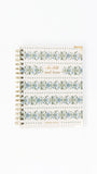 A spiral-bound notebook with a white cover decorated with blue and green floral patterns, dotted borders, and gold foil text reading 'Be still and know' and 'LEMONIN NOTES'.