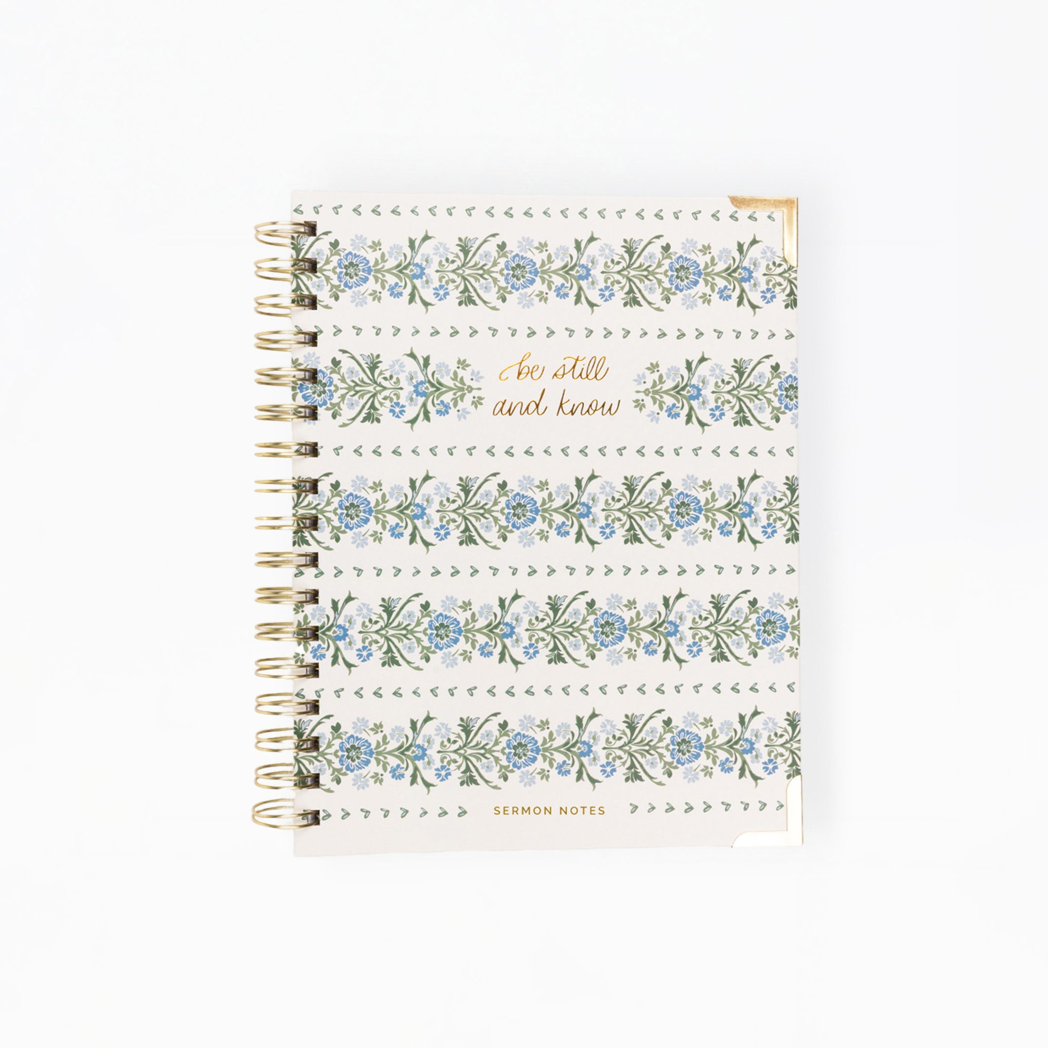 A spiral-bound notebook with a white cover decorated with blue and green floral patterns, dotted borders, and gold foil text reading 'Be still and know' and 'LEMONIN NOTES'.