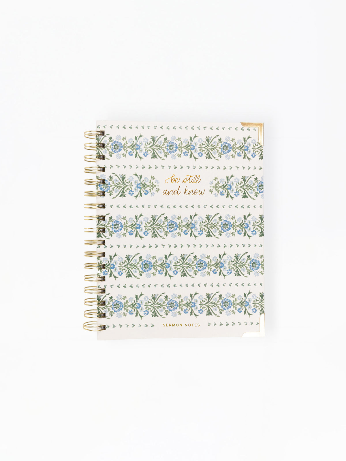A spiral-bound notebook with a white cover decorated with blue and green floral patterns, dotted borders, and gold foil text reading 'Be still and know' and 'LEMONIN NOTES'.