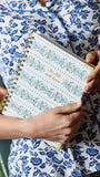 A person in a blue and white floral pajama shirt holds a spiral-bound planner or notebook with a white cover decorated with blue and green floral patterns and gold text reading 'See what you know'.