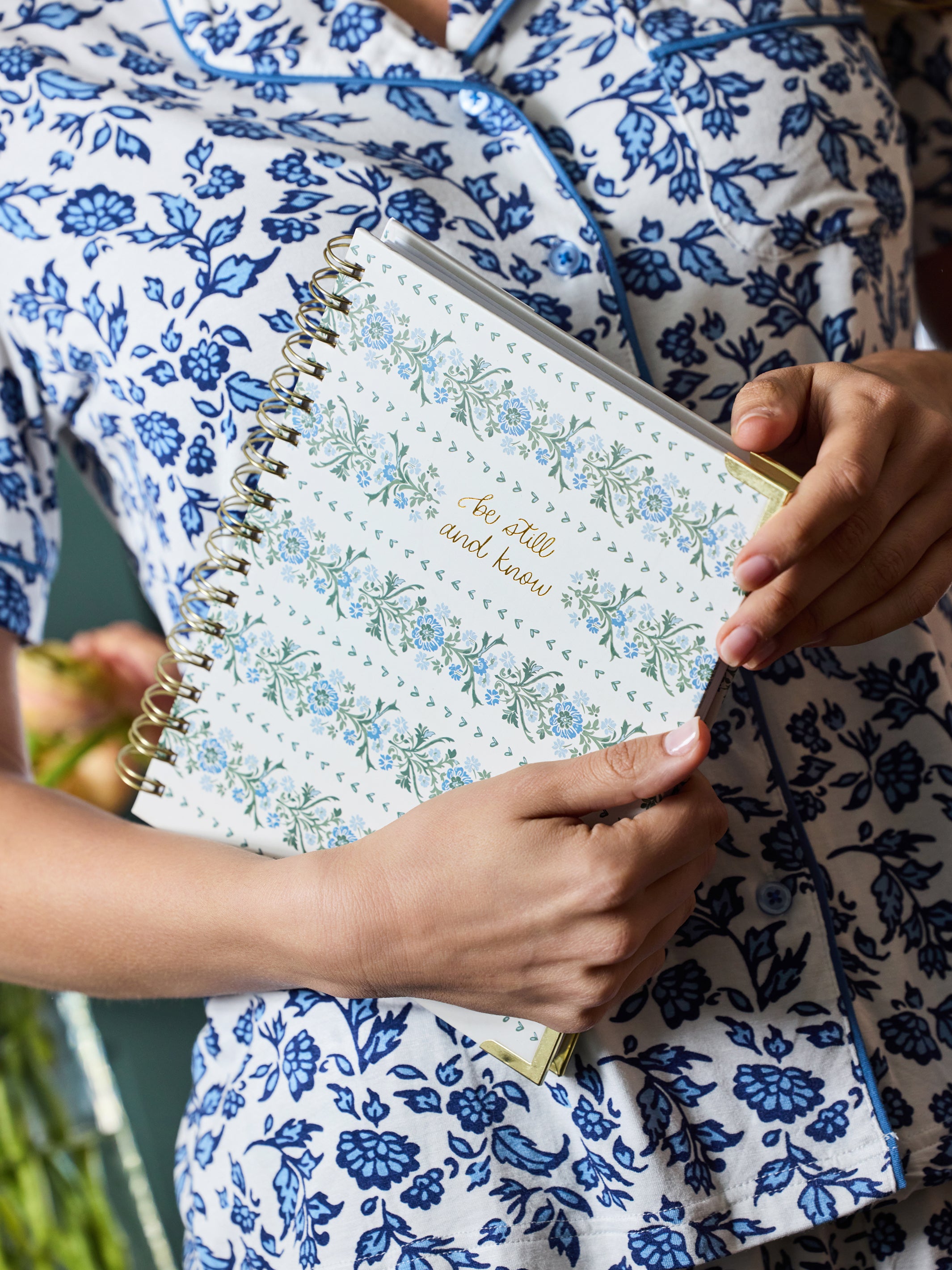 A person in a blue and white floral pajama shirt holds a spiral-bound planner or notebook with a white cover decorated with blue and green floral patterns and gold text reading 'See what you know'.