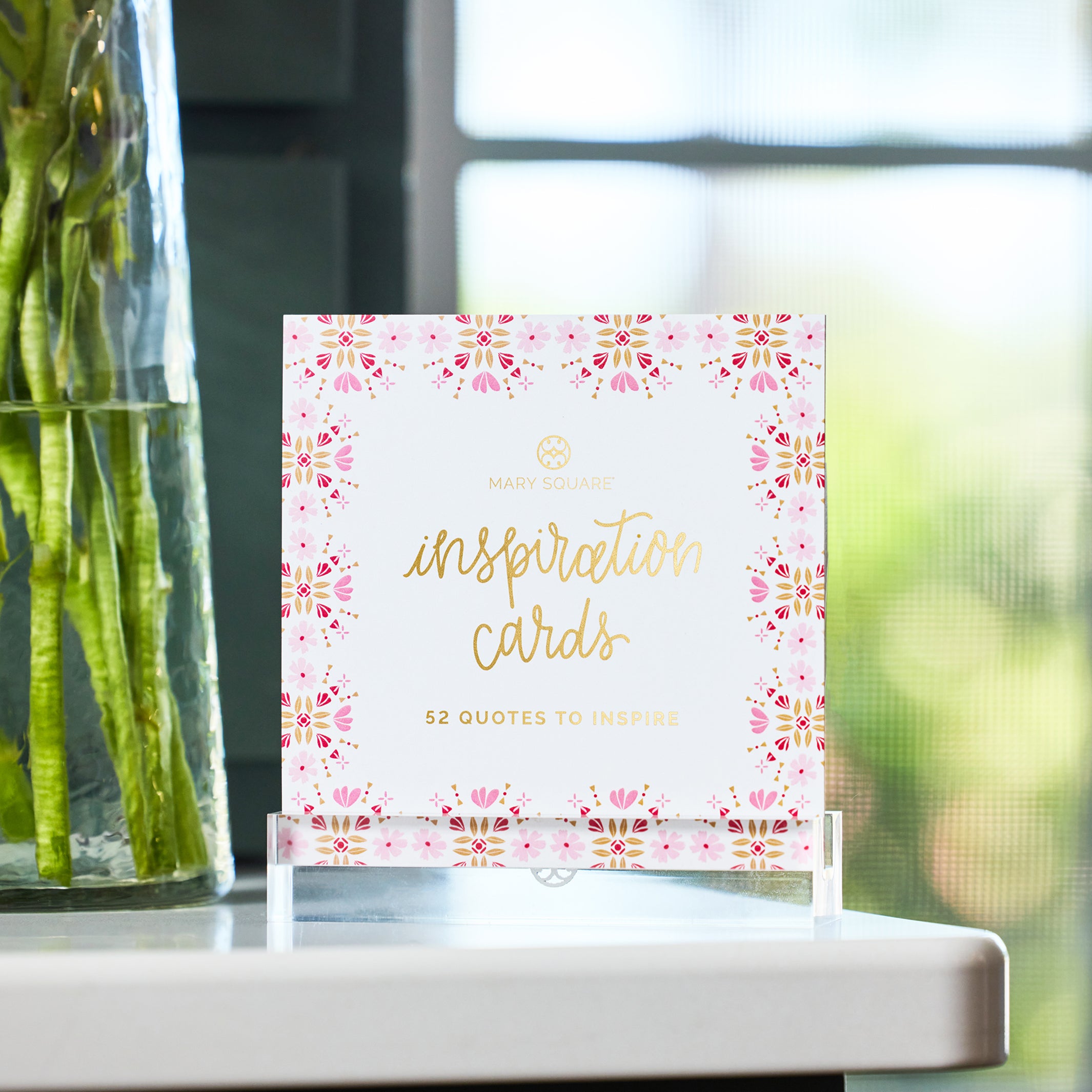 A small card stand holding a set of pink and gold 'inspiration cards' on a white tabletop, with a blurred vase of flowers and bright window light in the background.