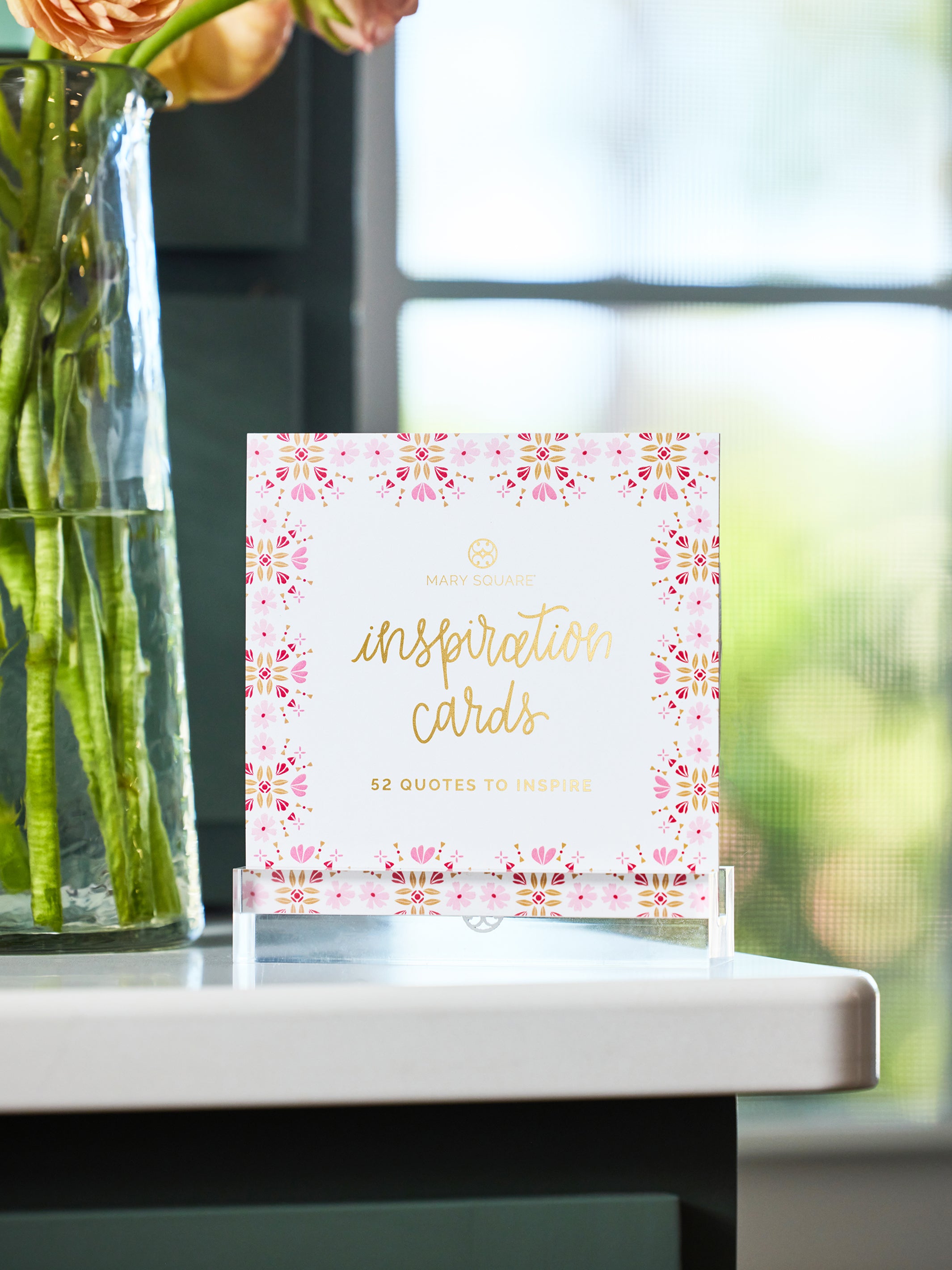 A small card stand holding a set of pink and gold 'inspiration cards' on a white tabletop, with a blurred vase of flowers and bright window light in the background.