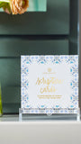 A boxed set of Mary Square scripture cards displayed on a clear acrylic stand on a shelf, with a blurred teal cabinet in the background and peach flowers in a glass vase at the left edge.
